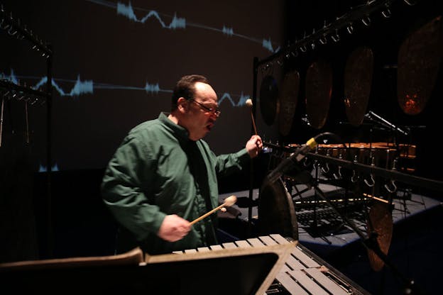 Winant animatedly plays percussion onstage. Bronze gongs hang to their right and behind them a screen projects blue sound waves.