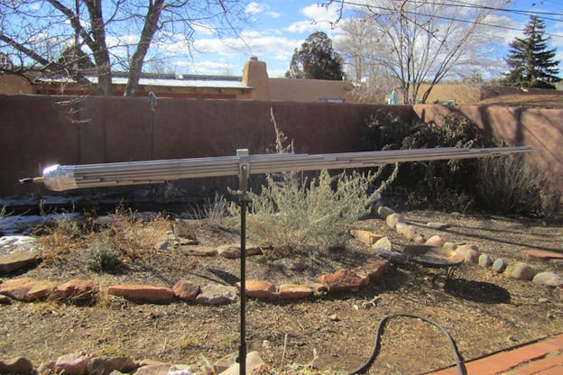 Horizontal metal structure shaped like adjoined tapered reeds in a tan-colored backyard filled with rocks, bushes, and remnants of snow. 