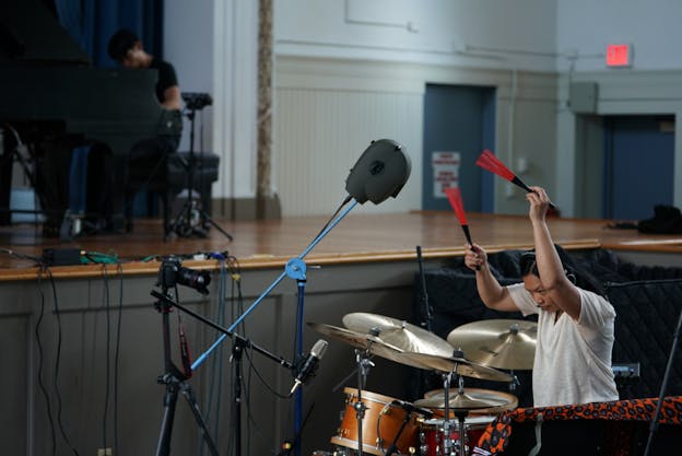 A photograph of Susie Ibarra performing with an orange drumset. She holds up two red drum brushes and faces the drumset with a concentrated expression. She wears a white t-shirt and headphones. In the background, there is an elevated stage with someone sitting and playing a grand piano. 