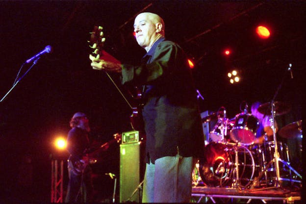 David First angles his body towards the camera and looks down at the guitar he is playing with a concentrated expression. He wears a black blazer and blue jeans. Behind him, there is another performer playing the guitar and someone else playing the drums. The entire scene is bathed in red and purple hued light. 