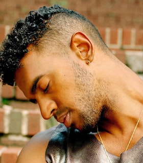 Portrait of Jonathan Gonzàlez at a side angle with his eyes closed. He is in front of a brick wall and wear a grey patterned tank top and a thin gold chain on his neck. 