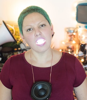 A portrait of Carmina Escobar in front of a desk of tangled cables, dressed in a burgundy t-shirt, a speaker's drum as a necklace and green hair, with a lighted lightbulb inside their open mouth.