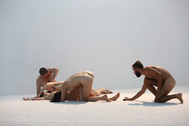 Four naked figures tangle together on a white floor, as a fourth naked person watches them.