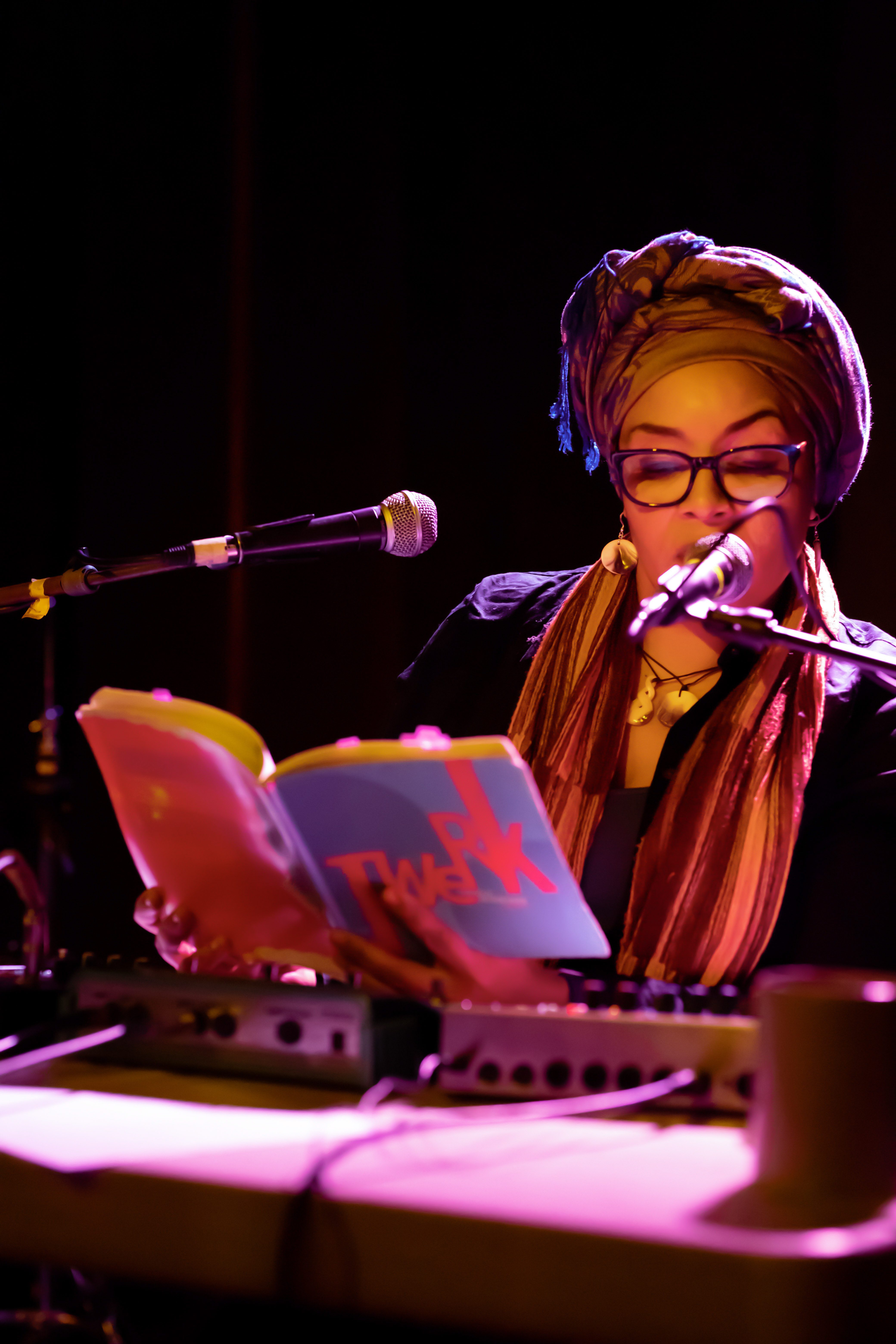 A figure sitting behind microphones and a booth, dressed in a headpiece and shawl with gold circular earrings and necklaces adorning them leans towards a microphone reading from a book with the title 'twerk.'