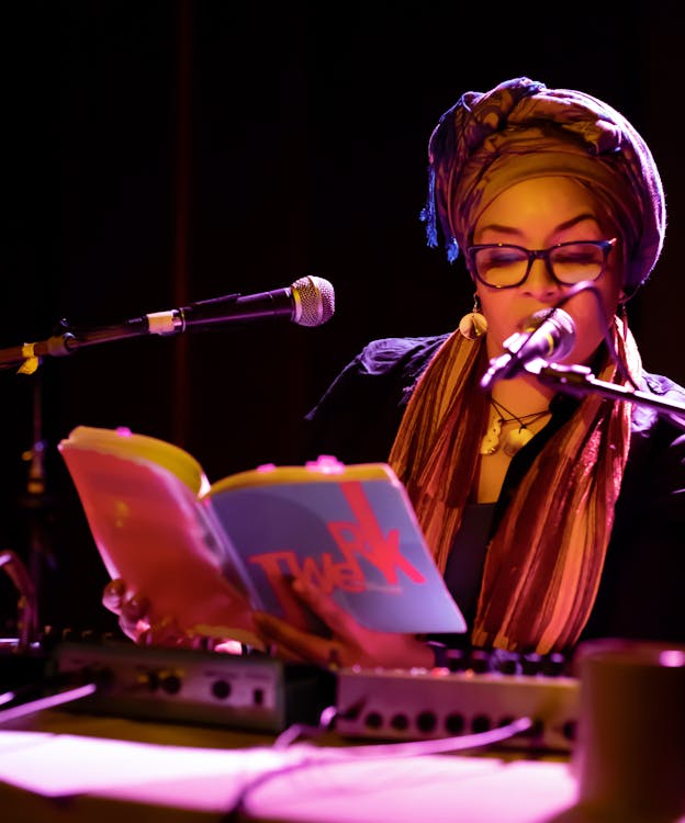 A figure sitting behind microphones and a booth, dressed in a headpiece and shawl with gold circular earrings and necklaces adorning them leans towards a microphone reading from a book with the title 'twerk.'