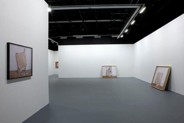Installation view of a multi-room gallery hosting framed photographs of wooden squares placed beside objects such as photographs on its walls and propped on its floor. 
