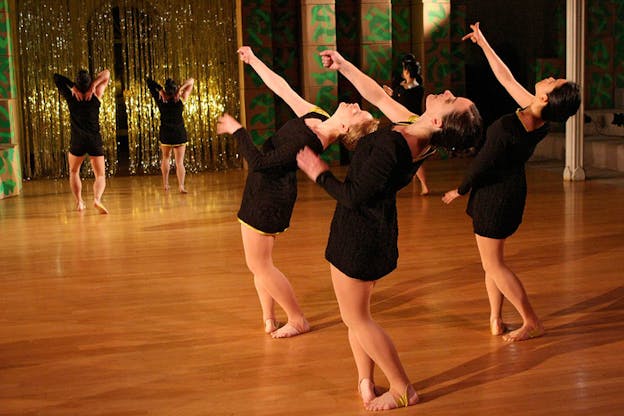 Three performers dressed in short black dresses and tan tights bend their knees and lean backwards with one elbow bent and one arm outstretched while in the distance other dancers strike poses in front of a wall partly decorated with gold tinsel. 
