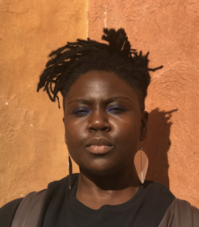 A portrait of Autumn Knight against a brushed orange background. She wears gold earrings shaped like leaves, a black shirt with brown leather detailing and blue eyeliner. She has short locked hair.