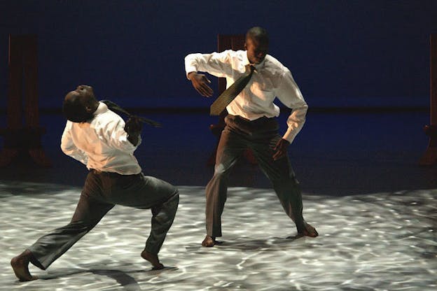 Two performers both wearing a tucked white shirt, a patterned necktie, and a pair of fitted pants stand under an ocean wave light projection. The one on the left performs a forward lunge while leaning their head back. The one on the right stares at the ground and slightly leans towards the other performer while holding their curled arms up. The dark background tinged with blue does not have a light projection.