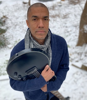 Morgan Craft looks directly at the camera, his face tilted up towards the camera above his eyeline. He stands in an out of focus snowy landscape, holding a black guitar by the body with the neck pointed towards the ground. He is wearing a black and white checkered scarf, and a navy corduroy jacket with black pants.
