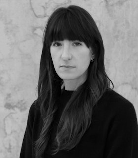 A black and white photo of Sam Contis turned at a three quarter angle facing the camera. She stands against a stone grey background, wearing a solid black sweater and small hoop earrings.