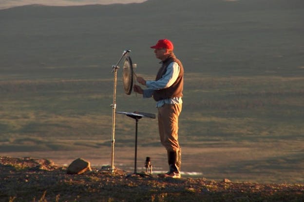 A figure dressed in tree trunk brown and wearing a red cap and glasses, holds two sticks with rounded ends that he hits a flat round plate with. He stands sideways on the middle of a grassy green plain.