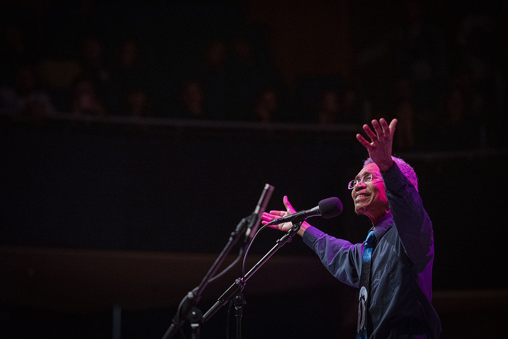 A performer with their hands extended upwards smiles in front of a stage microphone. Pink light illuminates on them.