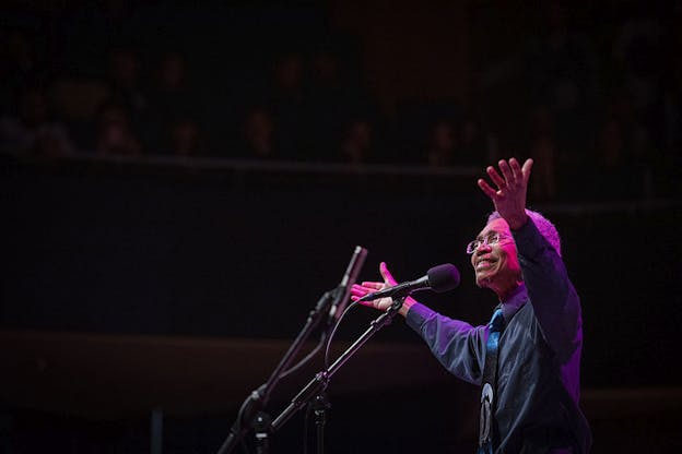 A performer with their hands extended upwards smiles in front of a stage microphone. Pink light illuminates on them.