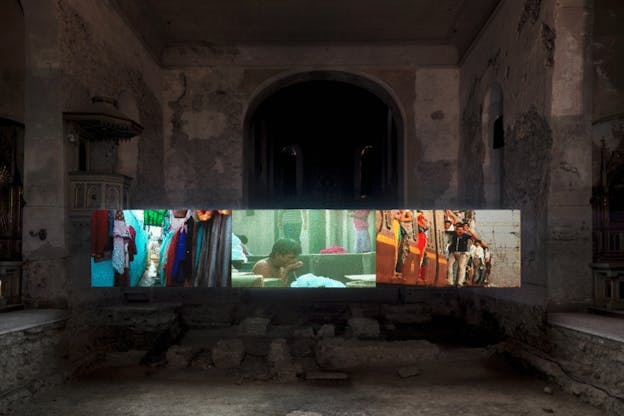 An installation image of a screen broken into thirds within a dark stone building. The images on the screen are colorfully hued and contrast sharply with the grey stone around them. On the left third of the screen, a thin street with clothing hanging from clotheslines is pictured. In the middle, there is an image of a man drinking water from his hand. On the right, there is an image of a crowded orange train with passengers leaning outwards. 