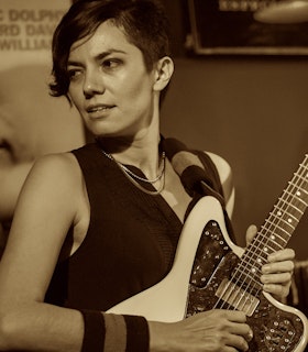 A sepia-toned photo of Ava Mendoza at a performance venue. She stands in sharp focus with their head turned towards the left while their body faces the right. Their eyes look to the left, they hold a white electric guitar with their right arm braced against the body, hand over the strings. Their other hand holds the lower neck of the guitar. They are wearing two chain necklaces, a black sleeveless v-neck blouse, and a thick, striped sweatband on their right wrist. 