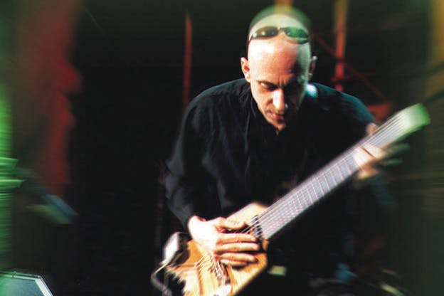 A slightly blurred photograph of a performance with Elliot Sharp playing guitar and leaning forwards towards the camera. He wears a dark shirt and thin black sunglasses. Bright red and green light emanates from the left and right edges of the photo.