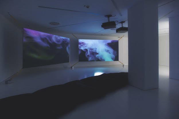 Photograph of a white gallery room with an elongated black sack material on the floor beginning from the left side of the image and ending to the other behind a column. Behind it in the nearby distance two projectors are set up on side by side screens showcasing a dark background with steam in colors of purple, aquamarine and green. 