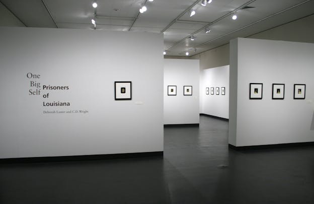 A photograph of an exhibition with several black framed images on different white walls. The title of the exhibition is in black font on the left side on a frame wall. 