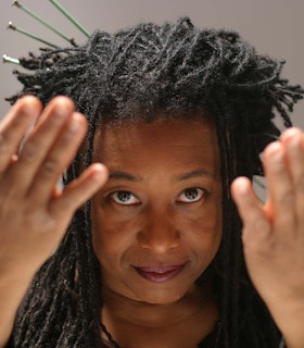 A close-up portrait of Pamela Z against a light grey background wearing purple lipstick and green stick pins in her hair. Her hands are raised by her face, palms facing up. 