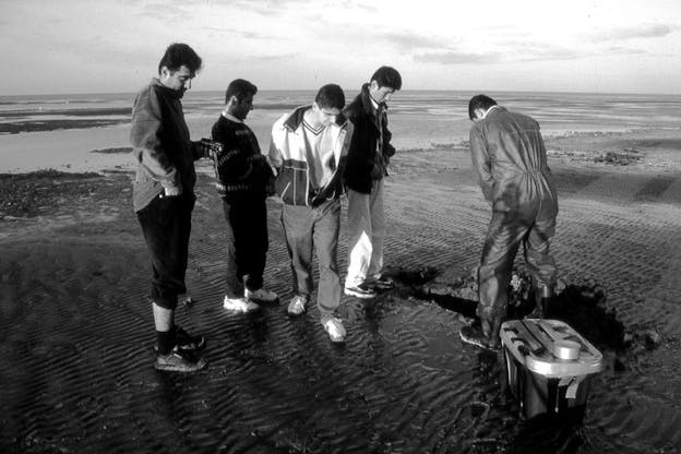 A black and white film still of five people clustered around a hole and looking into it. Beside the hole there is a small crate. They stand on an expanse of wetland.
