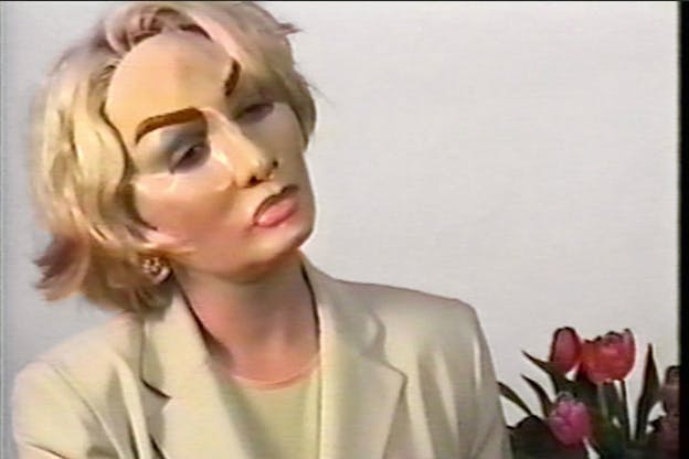 Blonde person dressed in a cream suit wearing a heavily maked-up mask. Red tulip flowers sit behind them, on a gray wall. 