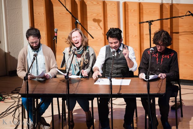 Four performers sitting behind a table with microphones in front of them reciting from papers. 