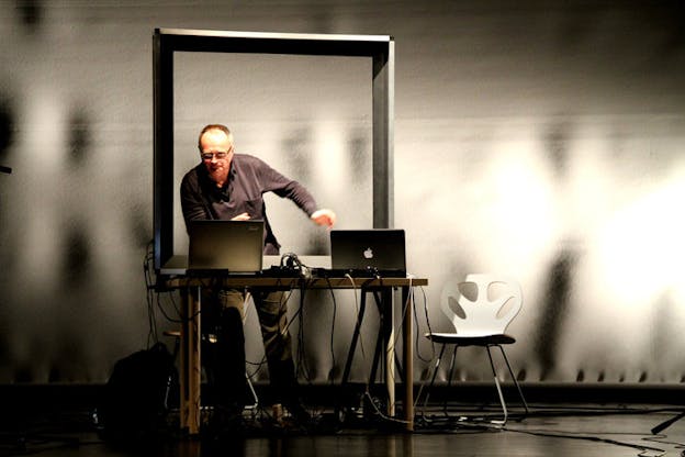 Knittel stands in front of two laptops in a gray space under a gray rectangle. He wears a dark purple shirt and appears deep in thought. Light dapples and coveres the entirety of the image in distinct light and shadow. 