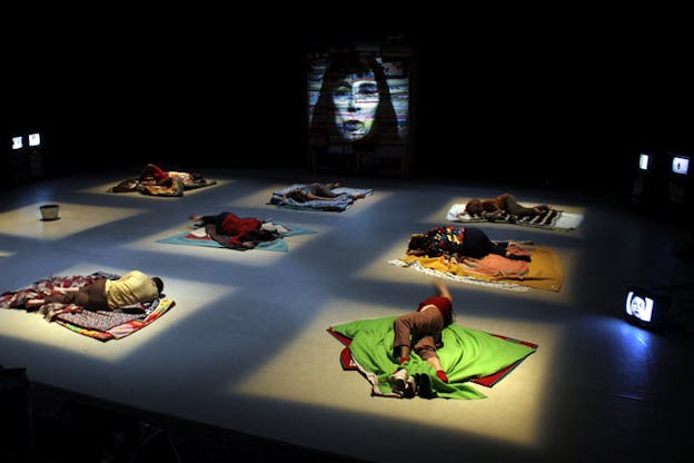 Performers on stage laying sideways on patterned covers on the ground, each separated and illuminated by square lights falling on them. Behind them the projection of a face with various lines of colors.