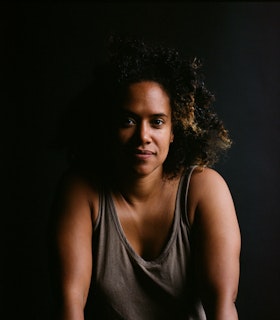 A portrait of Leslie Cuyjet wearing a grey tank top and facing the camera. The left side of her face is covered by shadow and the background of the image is black. 
