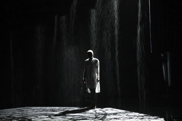 The silhouette of a figure in a white dress stands on a stage with rain pouring down. Their features are shadowed.