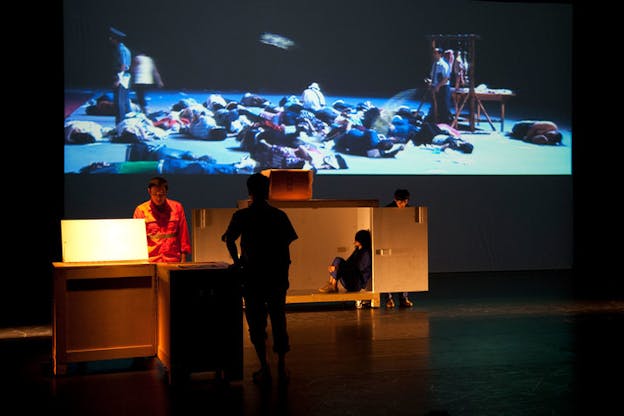 A performance still of two figures standing besides wooden tables. One figure has his back turned to the audience while the other faces the audience but looks downward. Behind them, another performer sits with their knees by their chest within a wooden box. Behind the door of this box, another performer stands, their body is hidden by the door so only their feet and their head, cast in a downward gaze, is visible. Behind all of this, there is a large blue hued projection of numerous people laying on the floor beside a rack with animal meat hanging from it. Two people are standing up. One of them is dressed like a police officer and the other is dressed in street clothes and directs a spraying hose towards the people on the ground. 