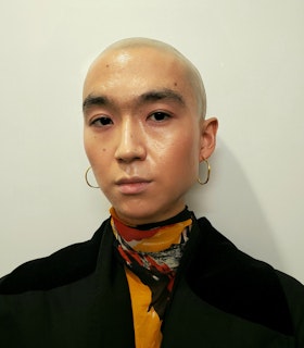 yuniya looks calmly into the camera. Her head is shaved and she is wearing medium hoop earrings, a black jacket, and a patterned scarf.
