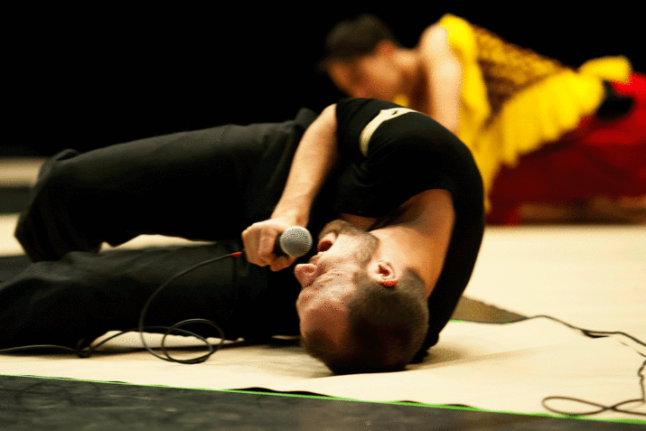 In-motion shot of a performer writhing on a white floor mat, singing into a microphone, their eyes squinted shut.