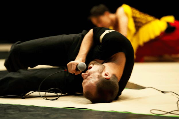 In-motion shot of a performer writhing on a white floor mat, singing into a microphone, their eyes squinted shut.