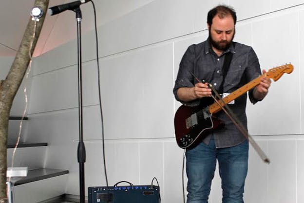 Layton in a gray button-up and jeans playing the guitar in a brightly lit white room. 