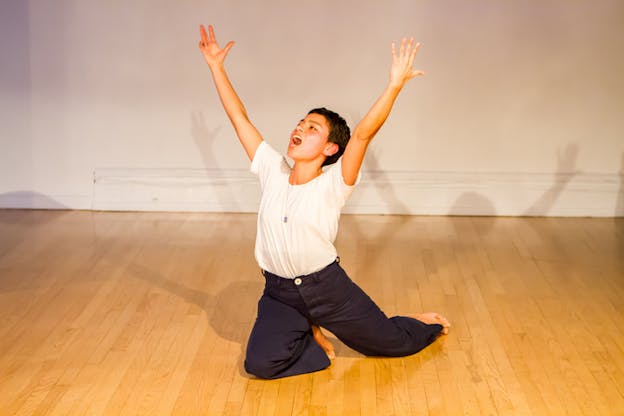 A performer in a white t-shirt and blue pants kneels on the floor with their arms upwards as they yell.