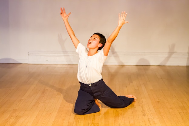 A performer in a white t-shirt and blue pants kneels on the floor with their arms upwards as they yell.