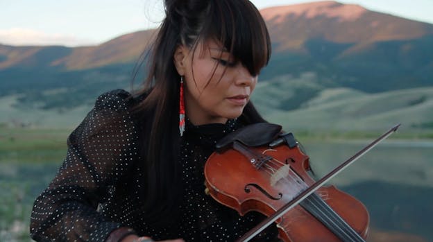 A video still of Laura Ortman performing with her violin against the backdrop of pink lit mountains. She wears a black and white polka dot blouse and long red beaded earrings.  Her eyes are closed and her head is tilted towards the violin. 