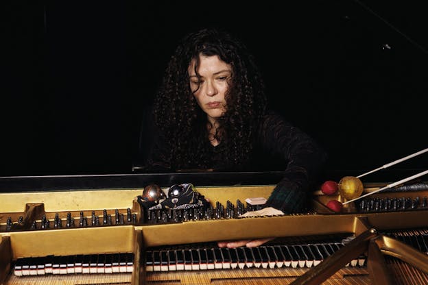 A figure clad in black with long dark brown curly hair behind a piano reaches towards the strings and dampers inside the open lid of the instrument.