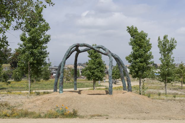 An organic sculpture of teal and brown stands outside, in the background there are patches of grass and trees.]