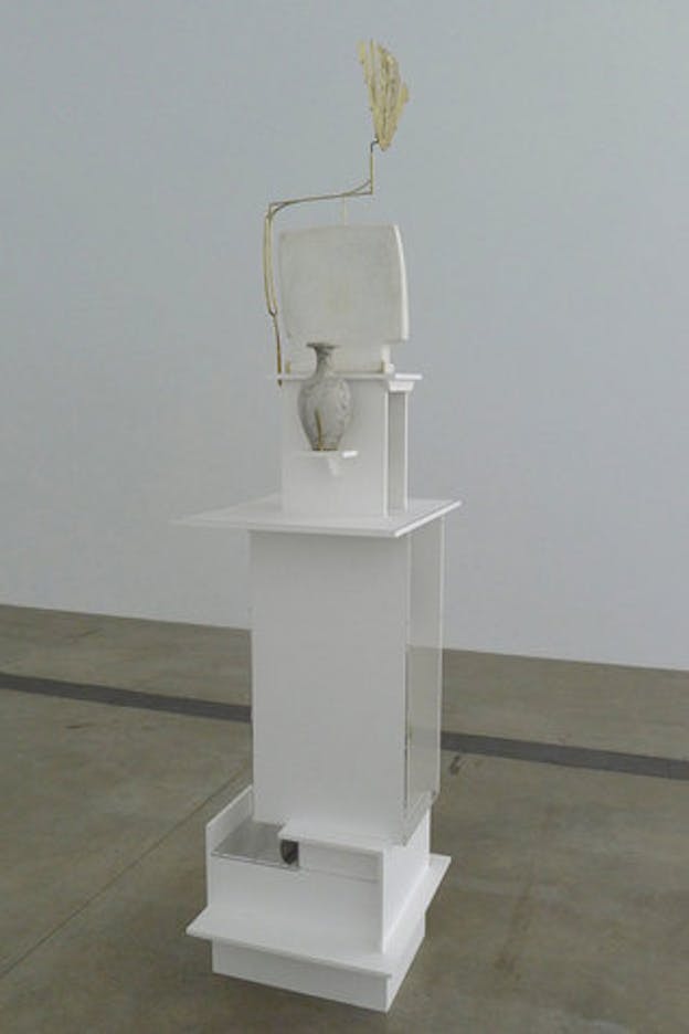 An installation image of a white shelf composed of numerous, differently sized, white boxes stacked atop each other. On one of the bottom boxes, there is a small horizontal sheet of a reflective, grey material. On another shelf, higher up, there is a grey and white marbled vase. On the top, there is a white stone rectangular sculpture surrounding by a thin golden rood which outlines the square and then continues upwards, ending in a leaf like topper. 