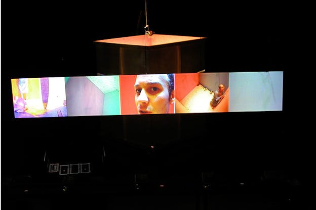 Projection of five aligned squares on dark surrounding. The projections hold two images of pink, yellow, gray and green colors to the left. In the middle projection a face and to its right a figure surrounded by orange walls. The last pictures is a white background.