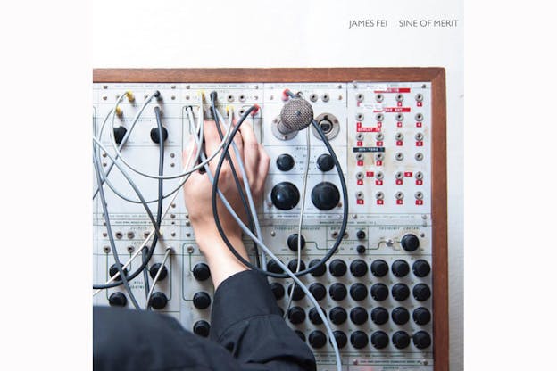 Close-up of a black-sleeved hand playing an analog electronic instrument, with rows of black knobs, a microphone, cables, and sockets marked with red letters. 