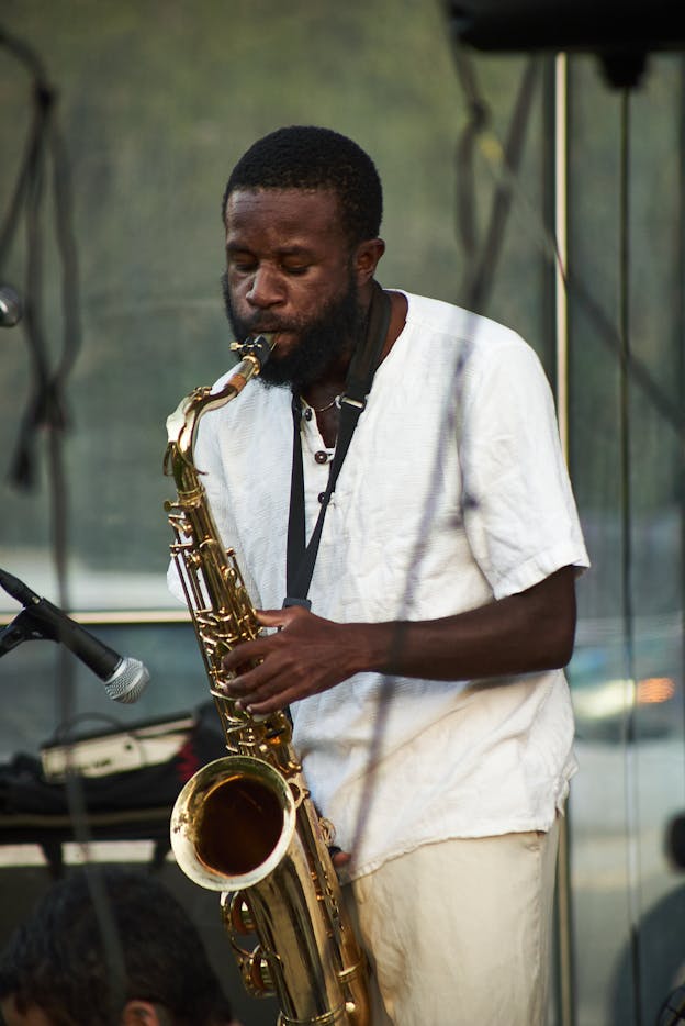 A photograph of JJJJJerome Ellis performing, closing his eyes and playing the tenor saxophone. He wears a textured white shirt and khaki pants. Around him, there is a microphone and other sound equipment. 