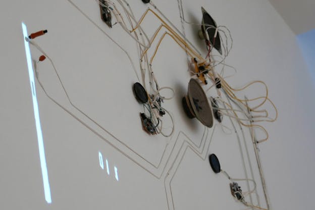 White and yellow cables stuck on a white wall connecting with multiple small electronic devices in circular and square shapes. At the end of the cables where two red plugs have been stuck, are projected a white line and numbers.