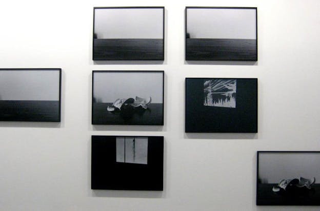 Close-up of a white wall hosting seven framed shadowy black and white photographs depicting a broken teacup, a white square resembling a window, and a dark strip against a white background resembling an ocean horizon. 
