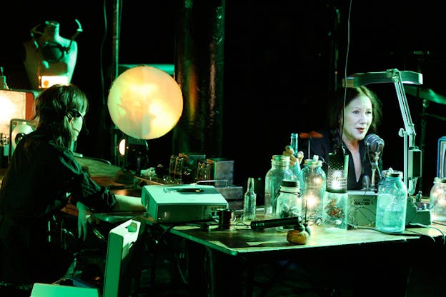 Black-Eyed-Susan sits at an emerald-green-hued desk, their face lit by an LED light speaking into a microphone while a person sits across from her, the back of their head visible.