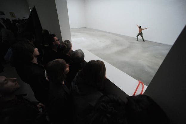 A shirtless performer on a gray stage opens their arms to the sides, as crowd watches from the top through a window cut in the wall.
