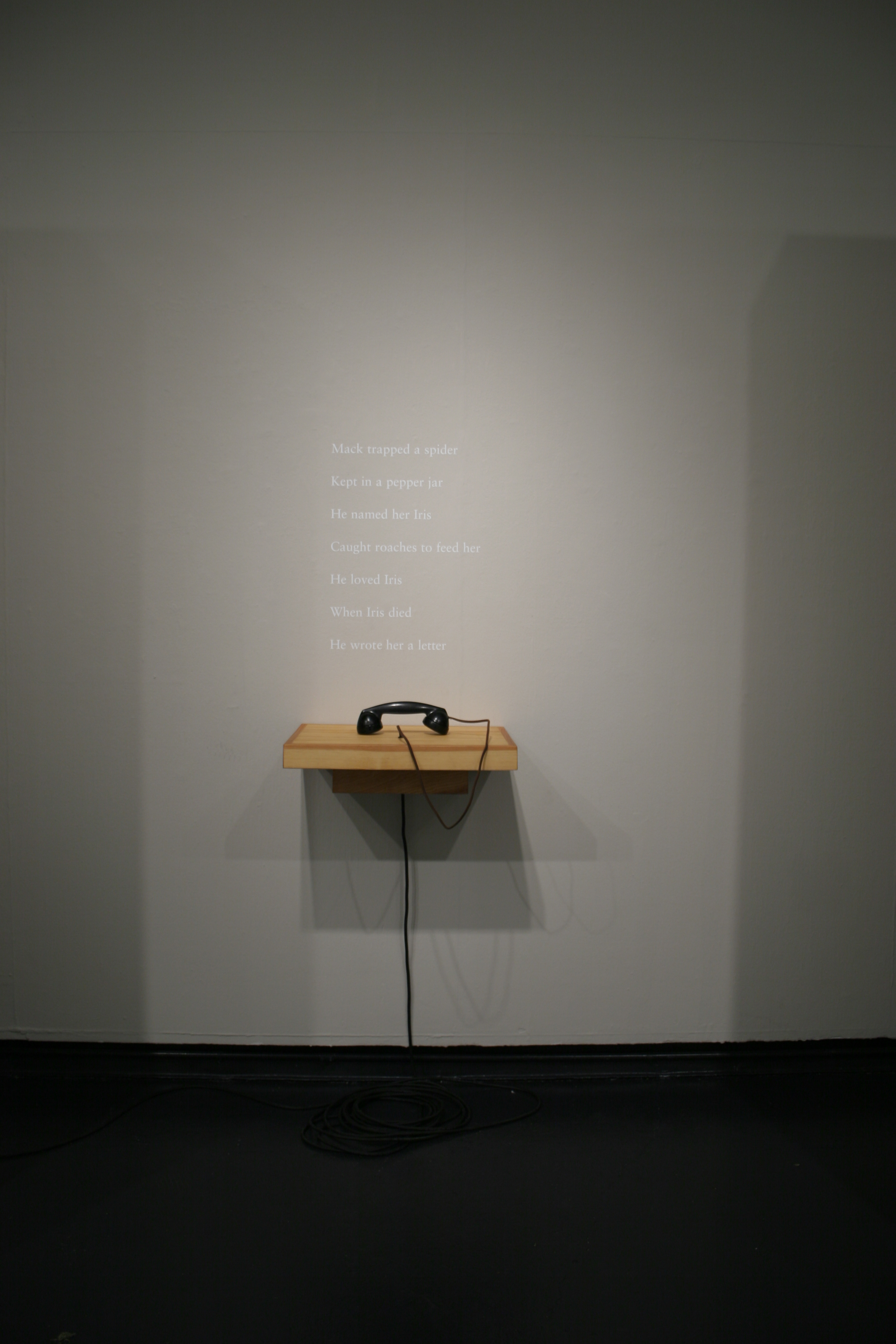 An exhibition photograph of a gray wall with a poem written on it in white text. Beneath the poem there is a wooden shelf with a black telephone on it.